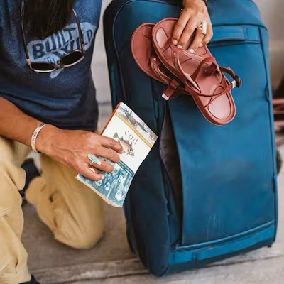 Person holding a sandal and book next to a blue suitcase.
