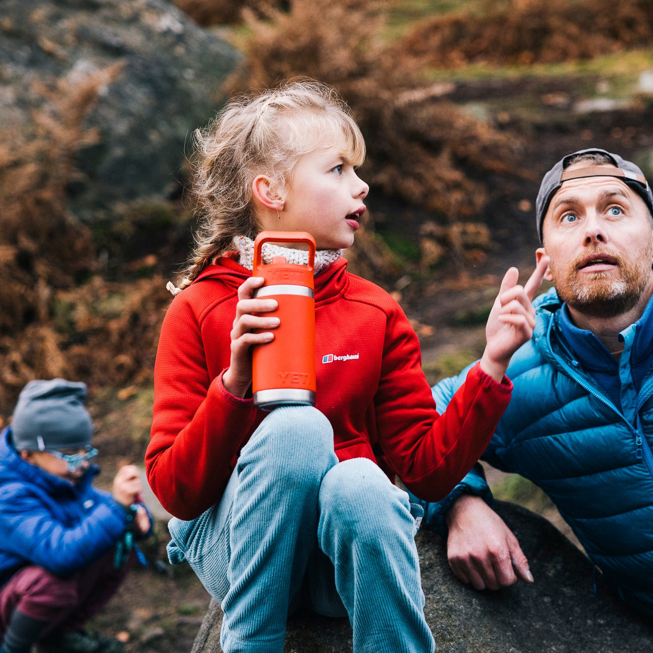Man and two children sitting on rocks outdoors, with one child holding a red YETI bottle.