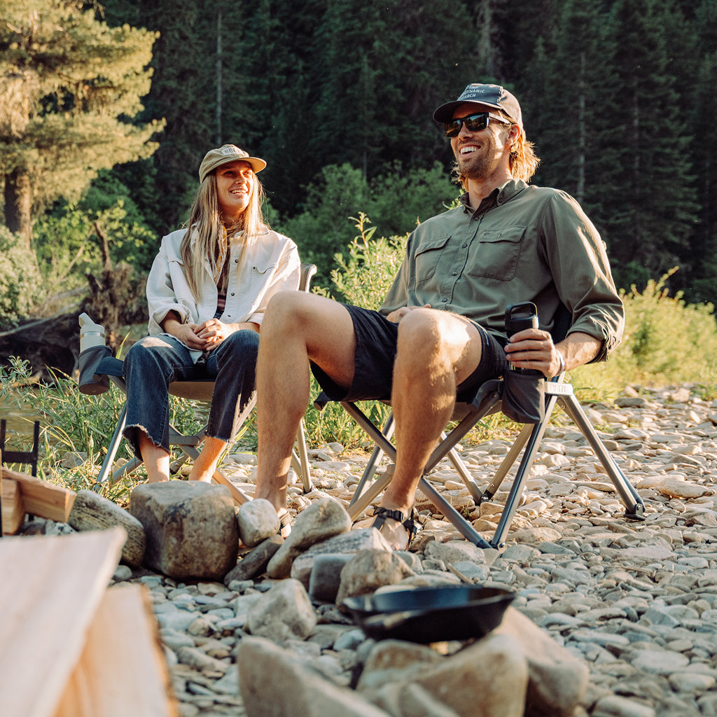 Two people sitting on YETI Field chairs by a campfire in a forest setting.