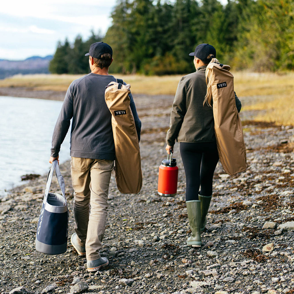 Two people walking along a rocky shoreline with outdoor gear, carrying YETI Field Chairs and YETI Camino, YETI Rambler jug.