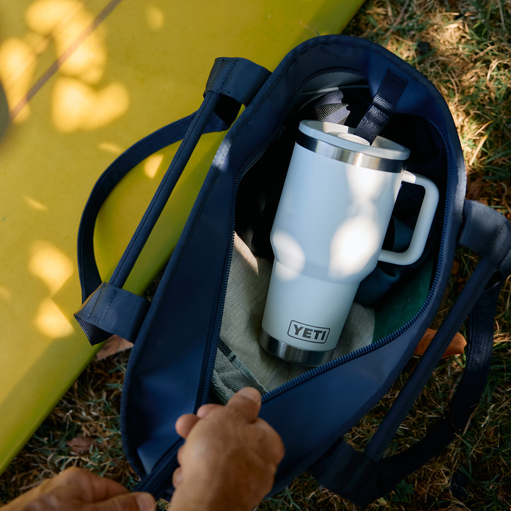 A navy blue tote bag is shown outdoors on grass, opened to reveal a white YETI insulated travel mug inside. A hand is visible holding the bag open, and a bright yellow board or blanket lies beside it.