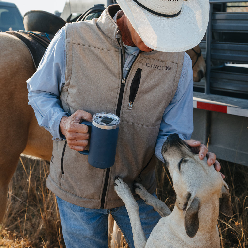 Man in cowboy hat and vest holding a blue mug with a dog in a field.