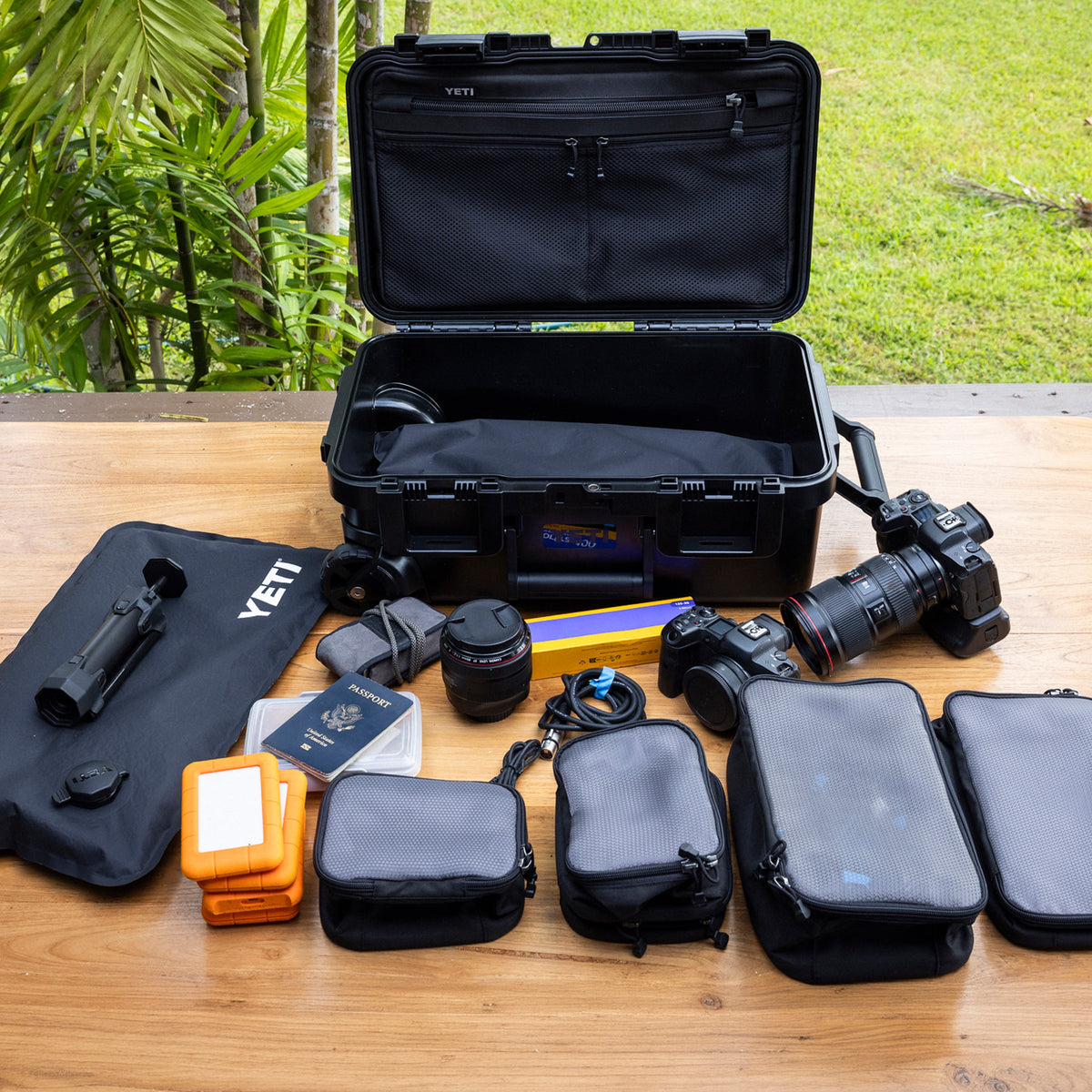 A YETI hard case opened on a wooden table, surrounded by camera gear including a DSLR camera, lens, accessories, and multiple black zippered pouches, with a garden background visible.