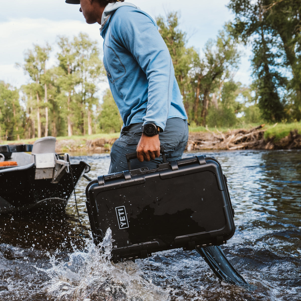 A man in a blue hoodie and jeans carries a black YETI hard case through splashing water next to a boat, with trees in the background.
