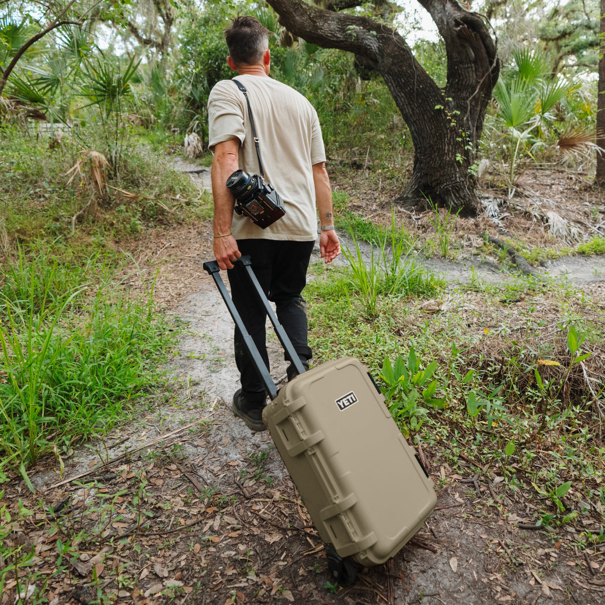 A man walking on a forest path pulling a tan hardshell rolling suitcase with a camera bag over his shoulder.